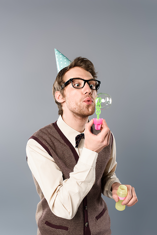 funny man in vintage clothing and party cap blowing soap bubbles on grey background
