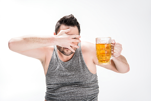 overweight man covering face with hand and holding glass of beer on white