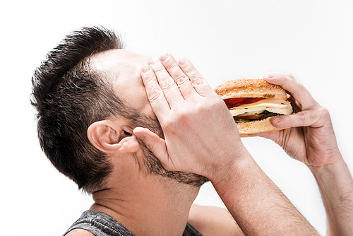 chubby bearded man covering face with hand while eating burger isolated on white