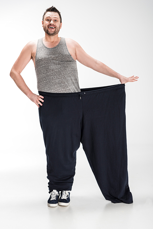 smiling overweight man holding oversize pants after weight loss on white