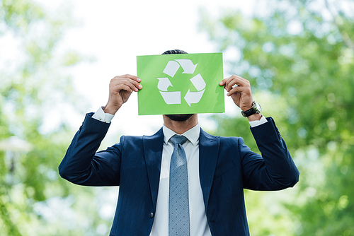 young man covering face with recycle sign card while standing in park