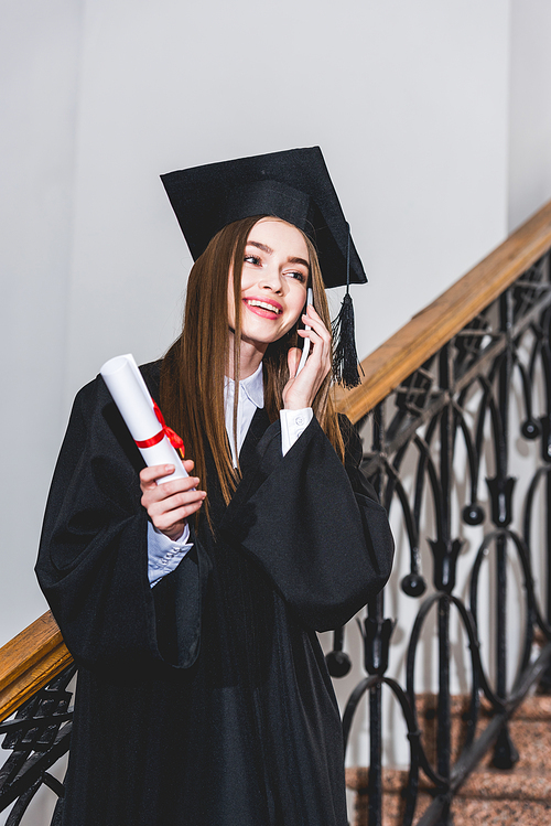 cheerful young woman holding diploma while talking on smartphone