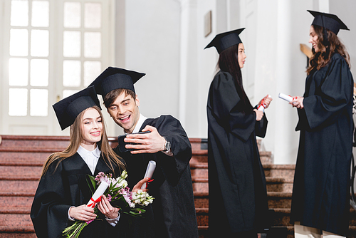 selective focus of handsome man and girl with flowers taking selfie near students in graduation gowns