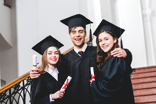 cheerful man in graduation cap hugging attractive girls while holding diploma