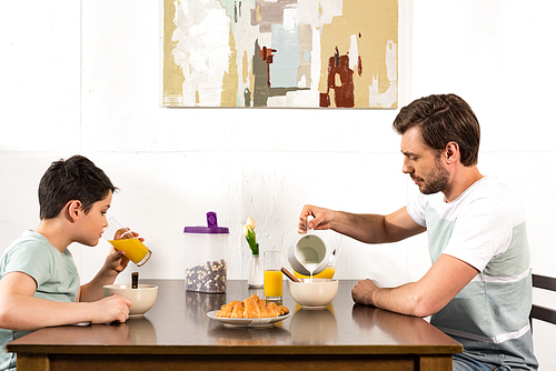 father pouring milk in bowl while sin drinking orange juice during breakfast