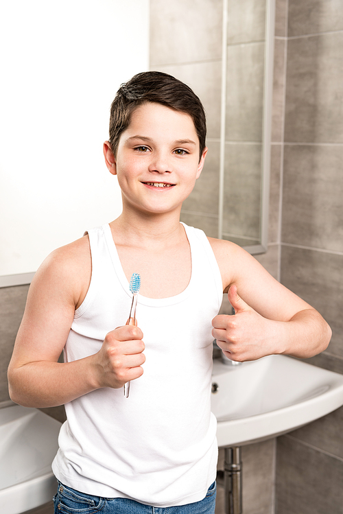 smiling boy holding toothbrush and showing thumb up in bathroom