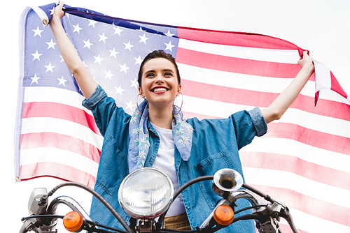 low angle view of beautiful girl sitting on scooter, holding American flag and smiling on sky background