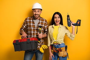 excited handywoman and handyman holding toolbox and electric drill on yellow