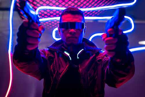 selective focus of armed mixed race cyberpunk player in futuristic glasses holding guns near neon lighting