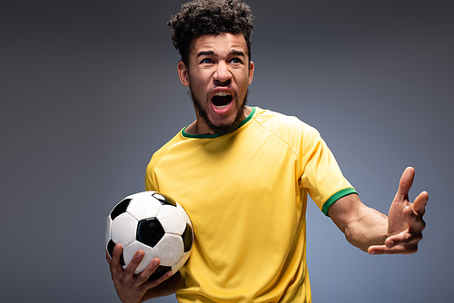 angry african american football fan in yellow t-shirt screaming and holding ball on grey