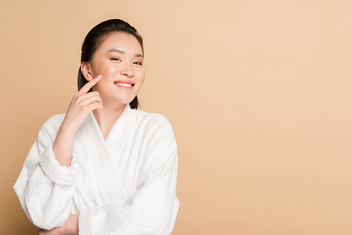 smiling beautiful asian woman in bathrobe touching face on beige background