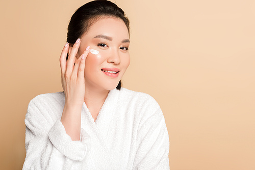 smiling beautiful asian woman in bathrobe applying face cream on beige background