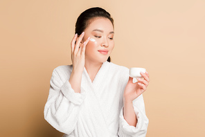 smiling beautiful asian woman in bathrobe applying face cream on beige background