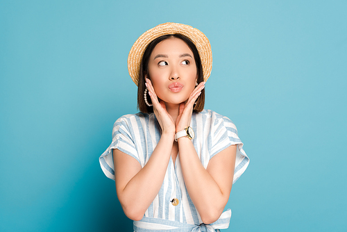 brunette asian girl in striped dress and straw hat touching face and pouting lips on blue background