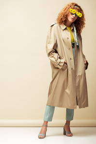 trendy redhead woman in trench coat and sunglasses with flowers standing with hands in pockets on beige