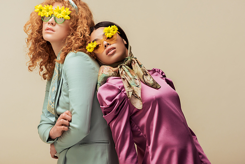 stylish african american girl and curly woman in sunglasses with flowers posing isolated on beige