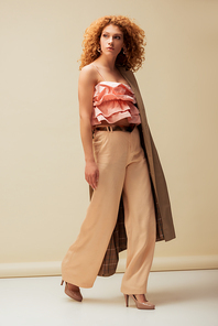 stylish redhead woman in ruffled top and trousers walking on beige