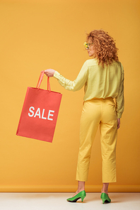 stylish redhead woman in sunglasses with flowers holding shopping bag with sale lettering on yellow