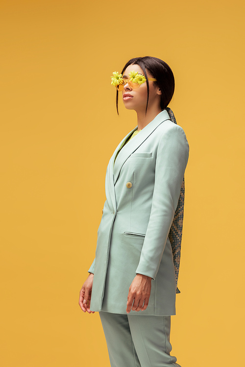 stylish african american girl in suit and sunglasses with flowers posing isolated on yellow