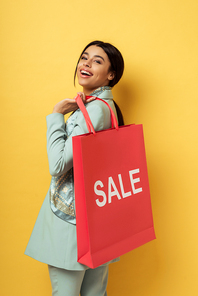 happy african american woman holding shopping bag with sale lettering and smiling on yellow