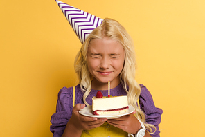 smiling kid holding plate with birthday cake and making wish on yellow background