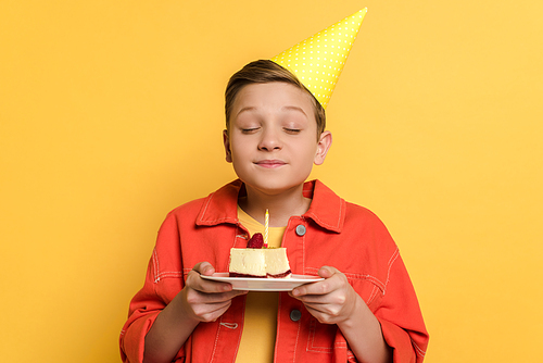 smiling kid with closed eyes making wish and holding plate with birthday cake on yellow background