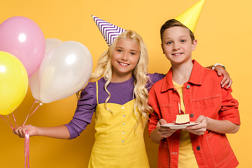 smiling kids with party caps holding balloons and plate with birthday cake on yellow background