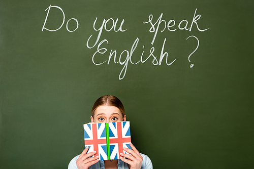 pretty girl with obscure face holding book with uk flag near chalkboard with do you speak English lettering