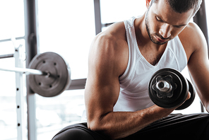 strong sportsman exercising with dumbbell while sitting in gym