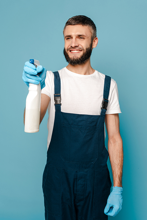 happy cleaner in uniform and rubber gloves holding spray detergent on blue background