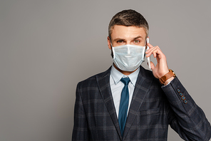 handsome businessman in medical mask talking on smartphone on grey background