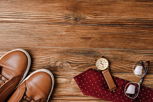 Top view of mens brown casual shoes, polka dots red tie, wristwatch and glasses on wooden background