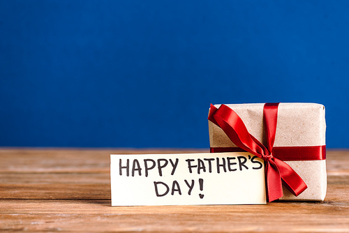selective focus of greeting card with lettering happy fathers day and gift box with red bow on wooden table isolated on blue
