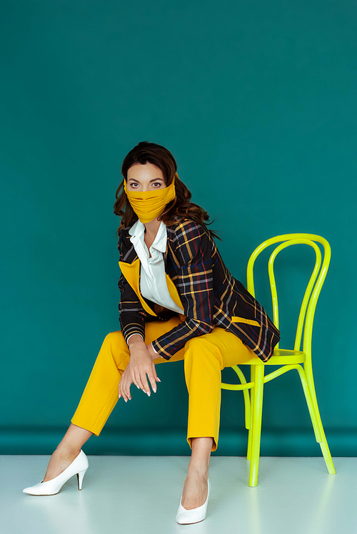 stylish woman in yellow mask and plaid blazer posing while sitting on chair on blue