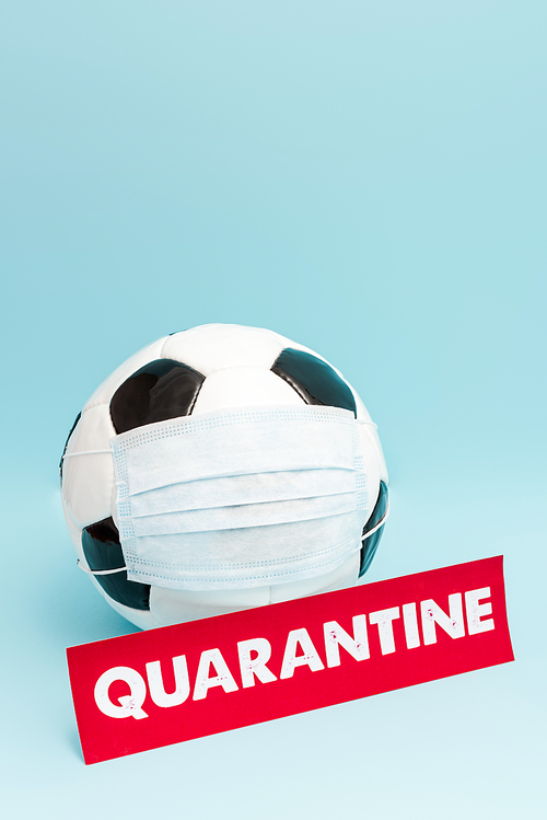 football in medical mask near paper with quarantine lettering on blue