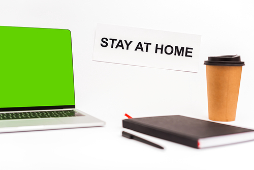 selective focus of laptop with green screen near paper cup, notebook, pen and paper with stay at home lettering isolated on white