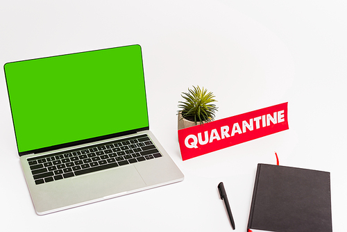 laptop with green screen near pen, notebook, plant and paper with quarantine lettering on white