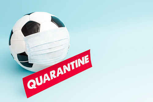 soccer ball in medical mask near paper with quarantine lettering on blue