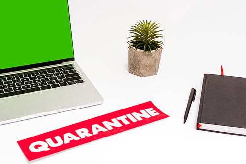 laptop with green screen near plant, pen, notebook and paper with quarantine lettering on white