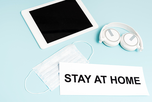 digital tablet with blank screen near headphones, medical mask and paper with stay at home lettering on blue