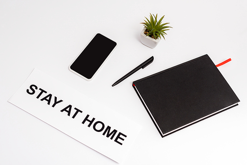 smartphone with blank screen near pen, notebook, plant and paper with stay at home lettering on white