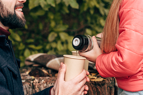 cropped shot of child pouring tea from thermos while smiling father holding paper cup in park