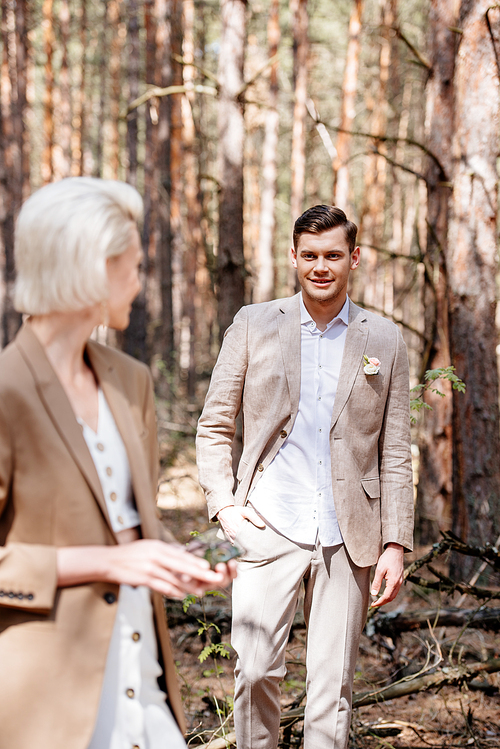 smiling bridegroom standing with hand in pocket and looking at bride in forest