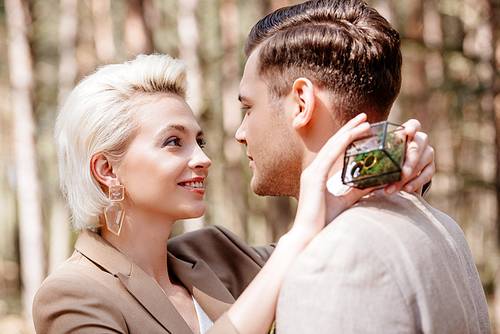 smiling blonde girl holding box with wedding rings and embracing bridegroom in forest