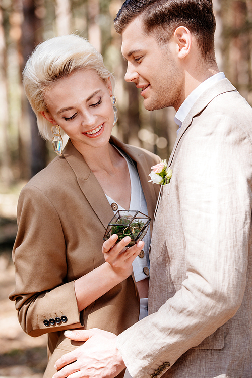 smiling bridegroom and bride holding box with wedding rings in forest