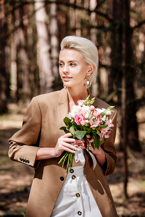 elegant blonde woman in earrings holding bouquet and looking away in forest