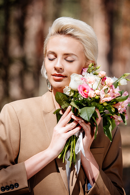 elegant young woman in brown jacket holding bouquet with closed eyes in forest