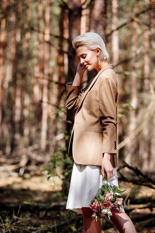 smiling blonde woman in jacket holding bouquet in forest