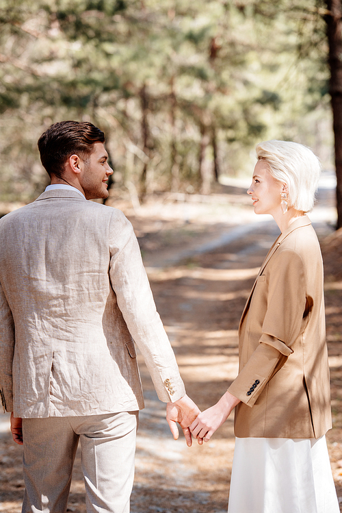 stylish smiling couple holding hands and looking at each other in forest