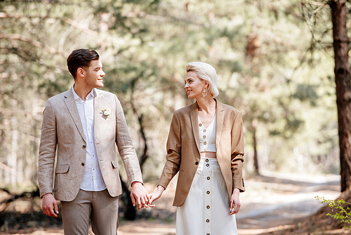 happy stylish couple holding hands and looking at each other in forest